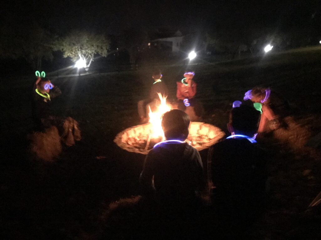 Boys sitting around a campfire with glow sticks on their heads. 