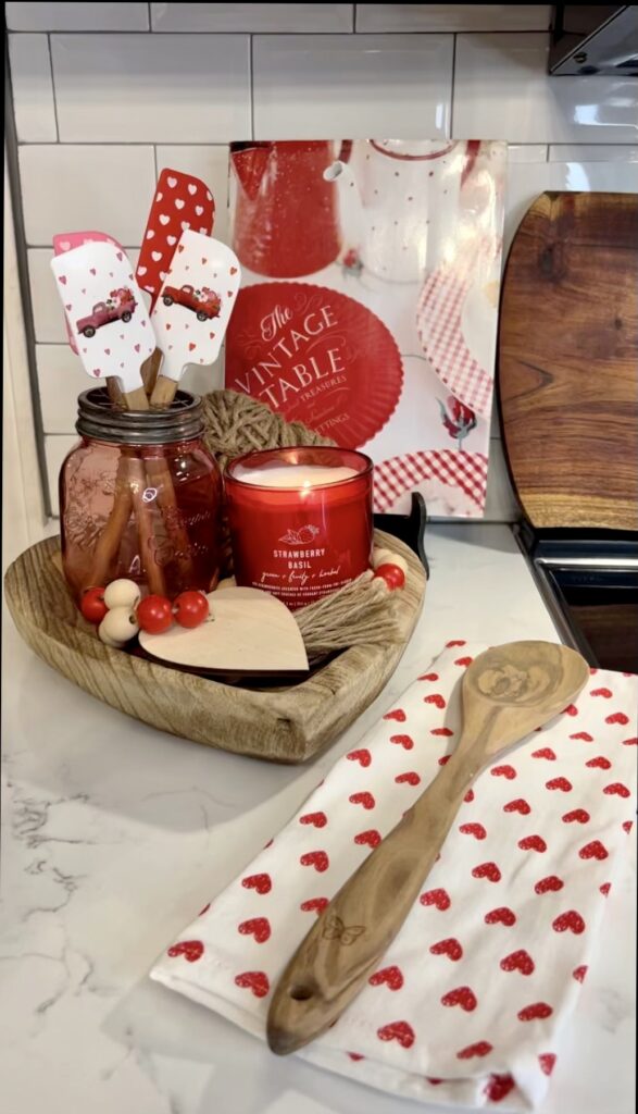 A picture of a cookbook and kitchen utensils with a candle and wood beads on a wood heart bowl. 